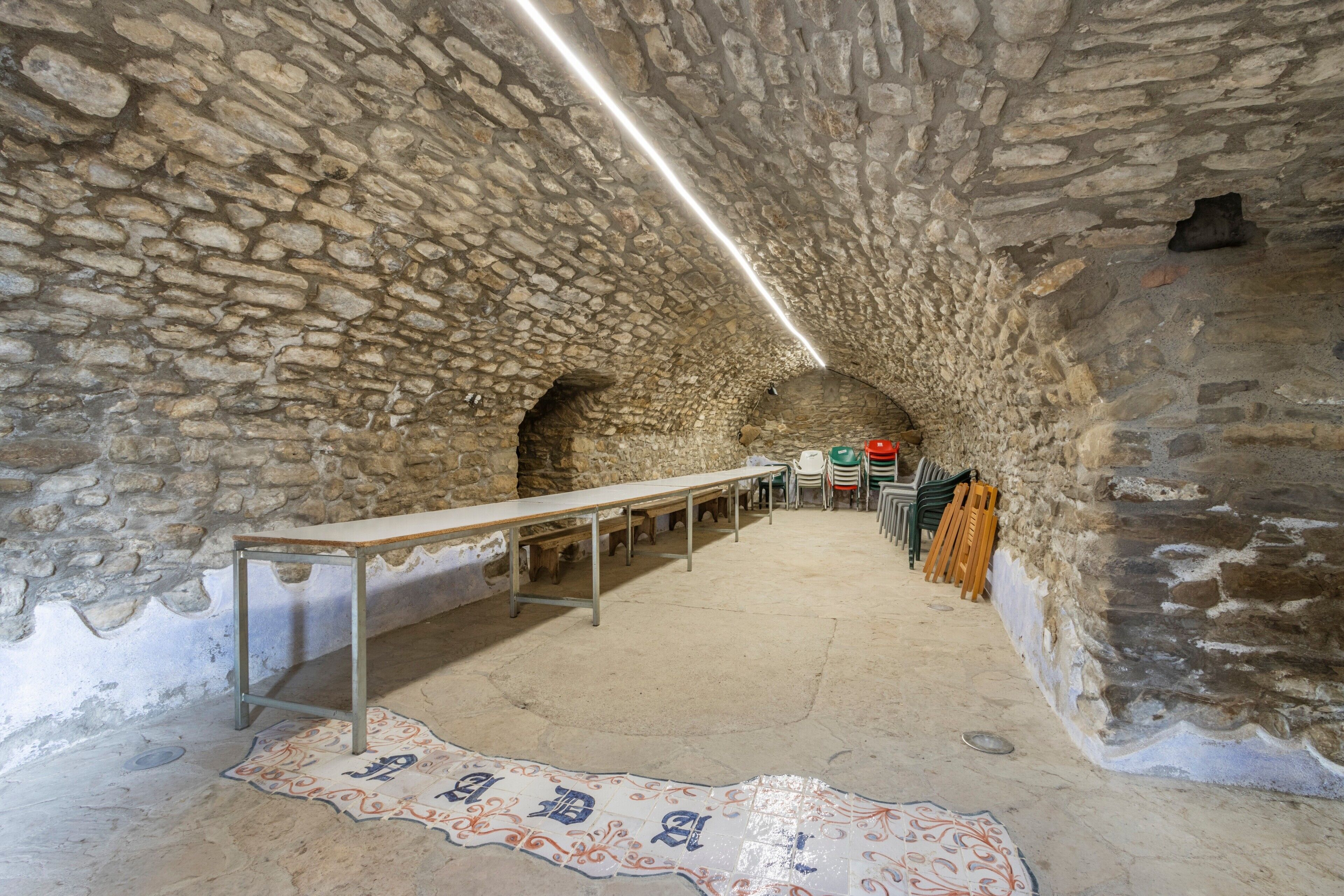 A stone-walled room with a long table and chairs, a patterned rug, and a ceiling light.