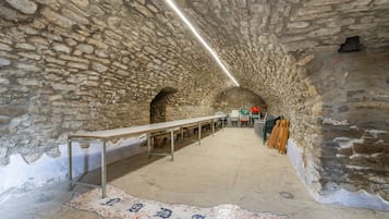 A stone-walled room with a long table and chairs, a patterned rug, and a ceiling light.