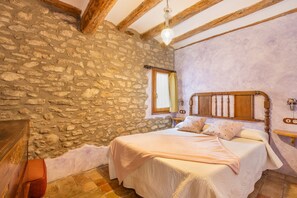 A bedroom with a stone wall, wooden beams, a bed with pillows, a nightstand, and a hanging light fixture.