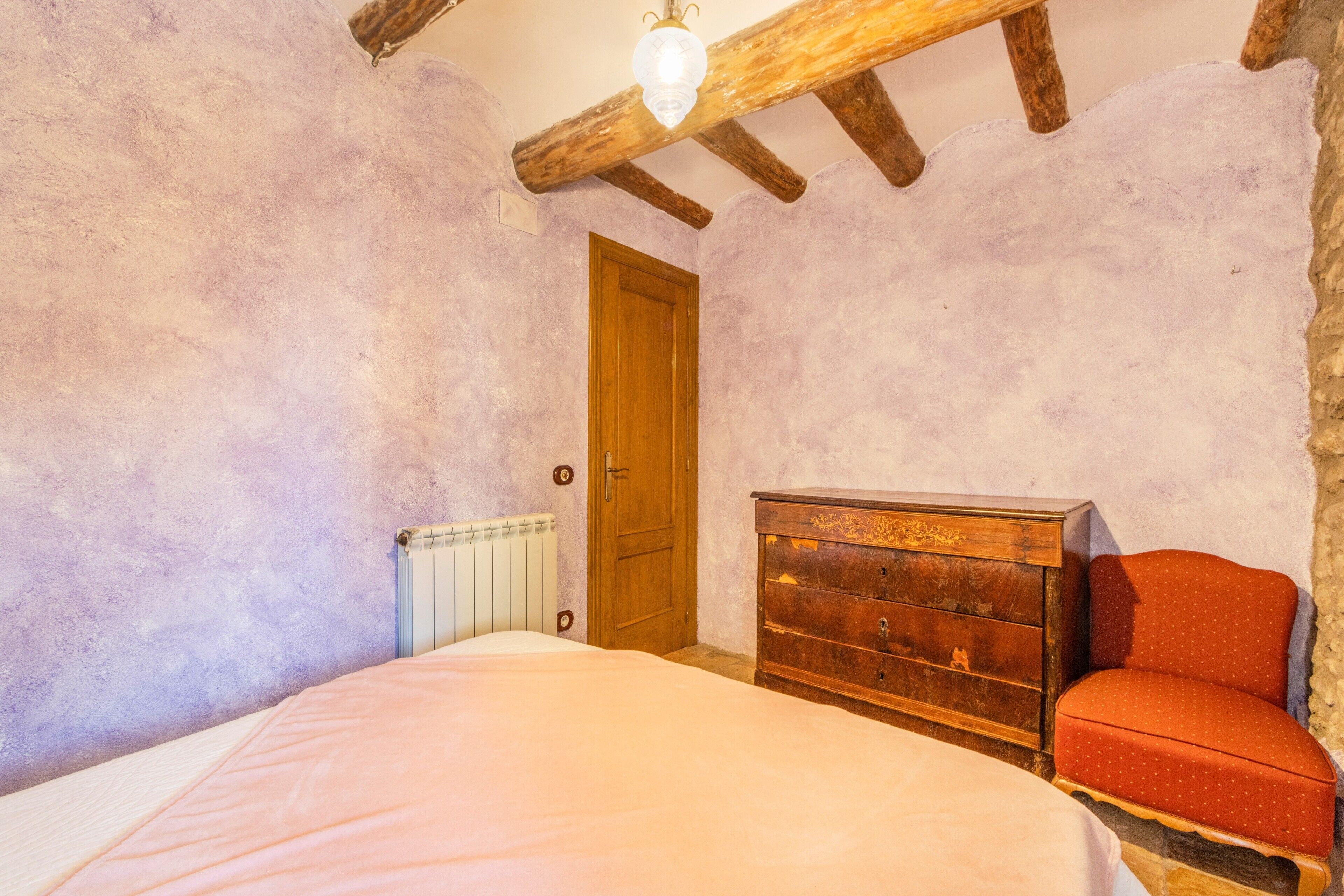 A bedroom with a wooden ceiling, a wooden dresser, a red chair, a bed with a white bedspread, and a radiator.