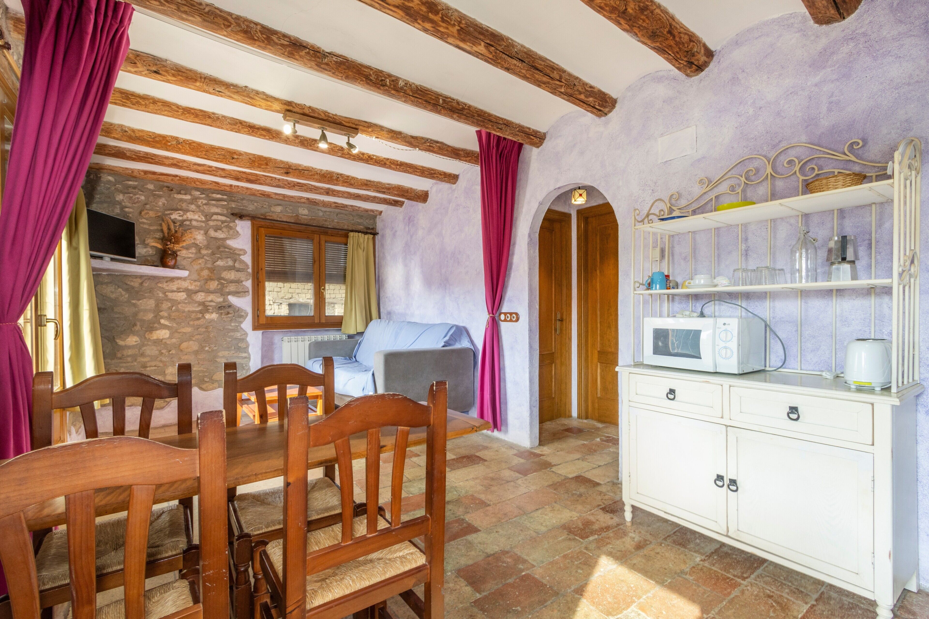 Una habitación rústica con vigas de madera a la vista, pared de piedra, mesa de comedor de madera con sillas, un armario blanco con electrodomésticos de cocina y un sofá acogedor.