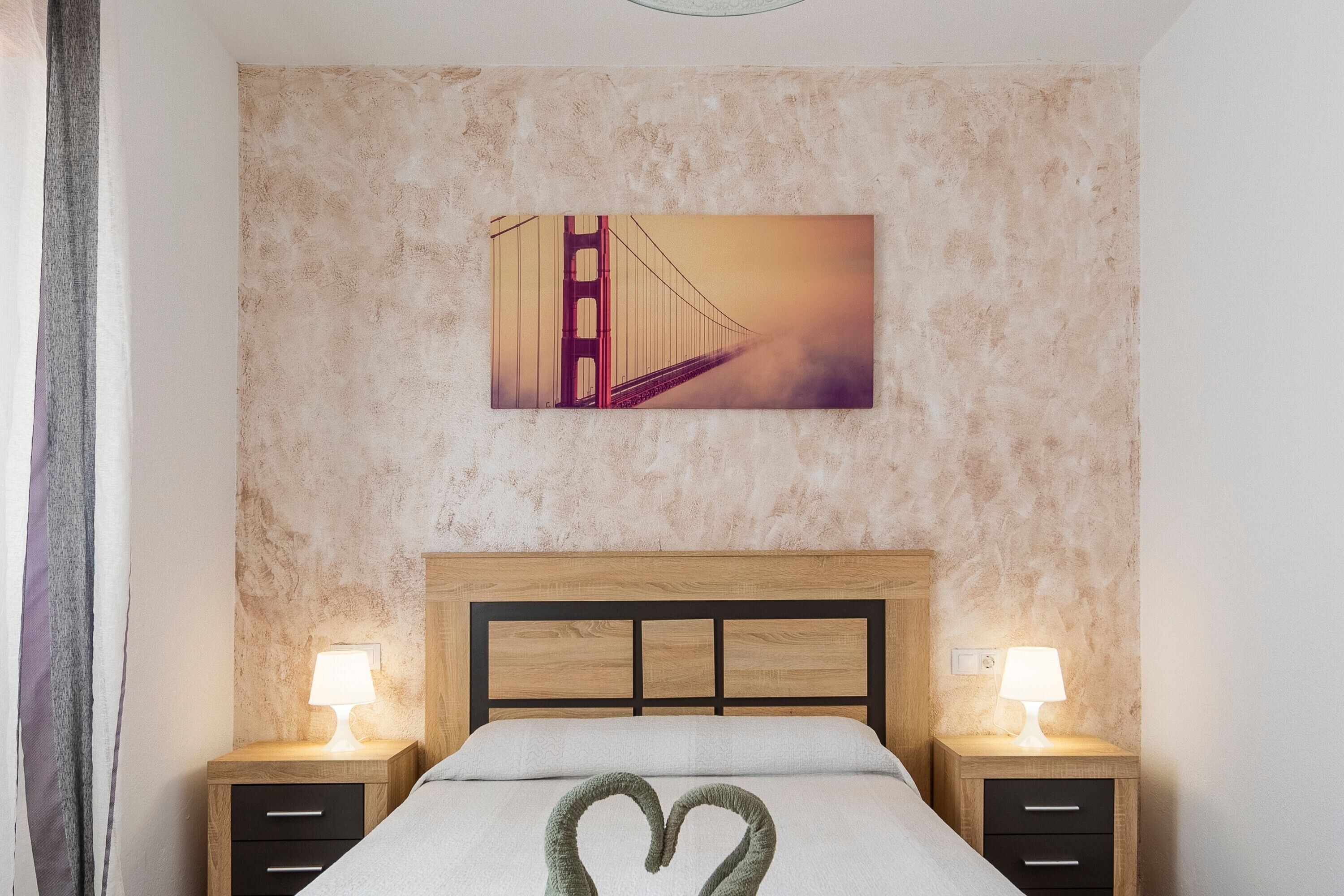 Une chambre d’hôtel avec un lit, deux tables de chevet avec des lampes, un tableau représentant le Golden Gate Bridge accroché au mur, et une décoration en forme de cœur sur le lit.