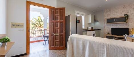 A hotel room with a bed, a TV, a kitchen area, and a view of palm trees through an open door.