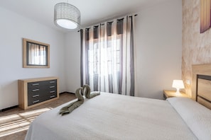 Una camera da letto con un letto, una cassettiera, un comodino con lampada, una finestra con tende e un quadro appeso al muro.