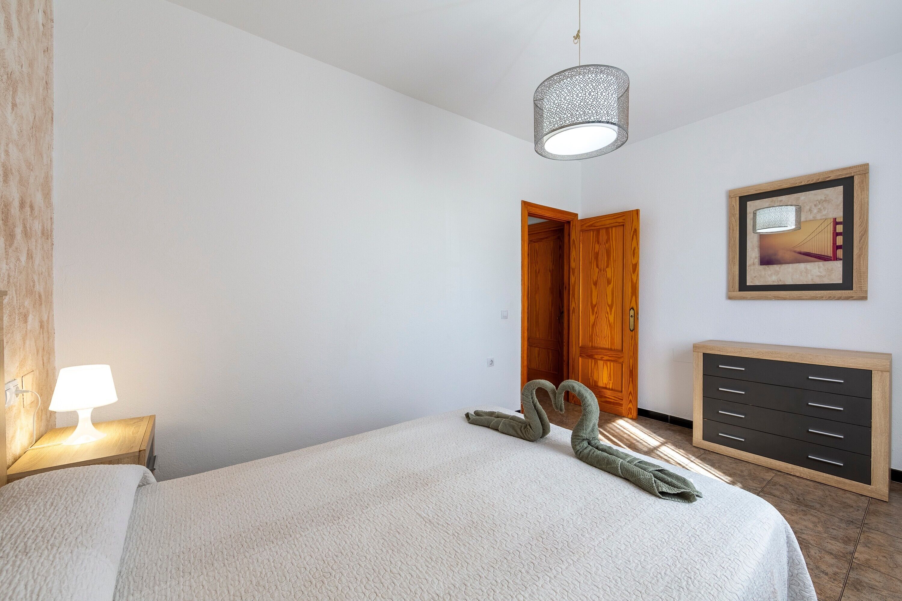 A bedroom with a bed, a bedside lamp, a dresser, a door, and a framed picture on the wall.