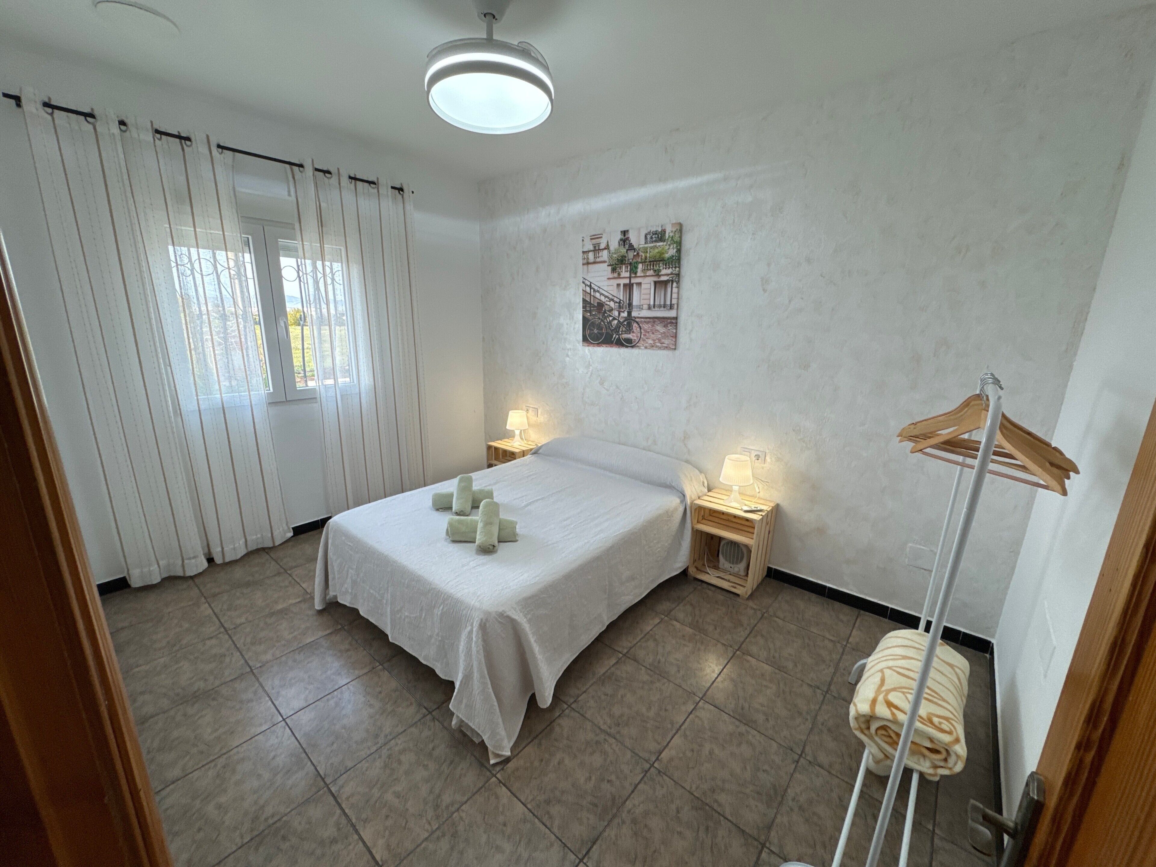 A bedroom with a bed, two bedside tables with lamps, a window with curtains, a wall-mounted picture, and a wooden drying rack.