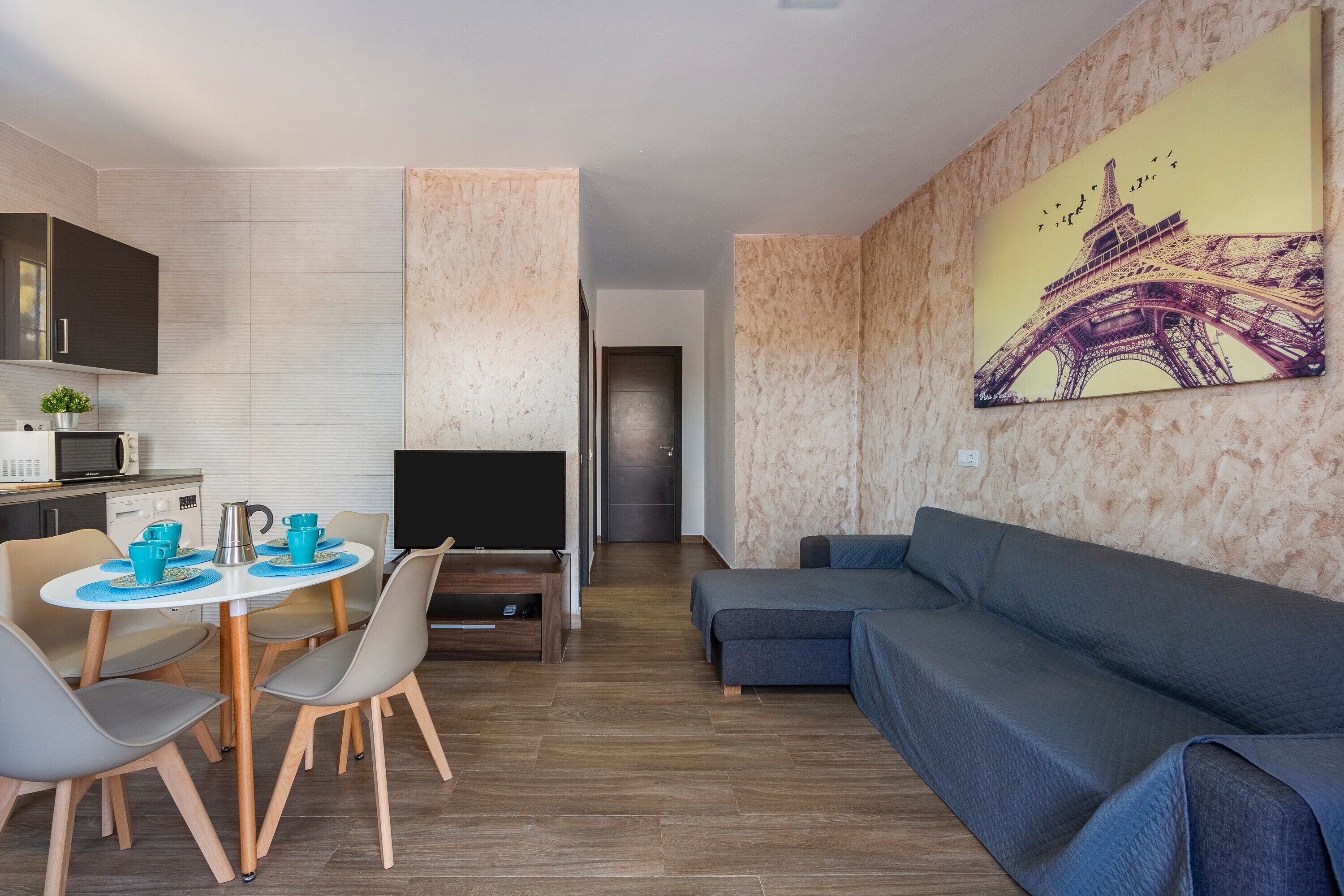 A modern living room with a sofa, a dining table with chairs, a TV, and a kitchen area in the background.