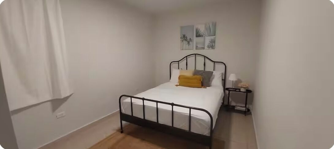 Ein kleines Zimmer mit einem Einzelbett, Metallgestell, weißer Bettwäsche und einem kleinen Nachttisch mit Lampe.