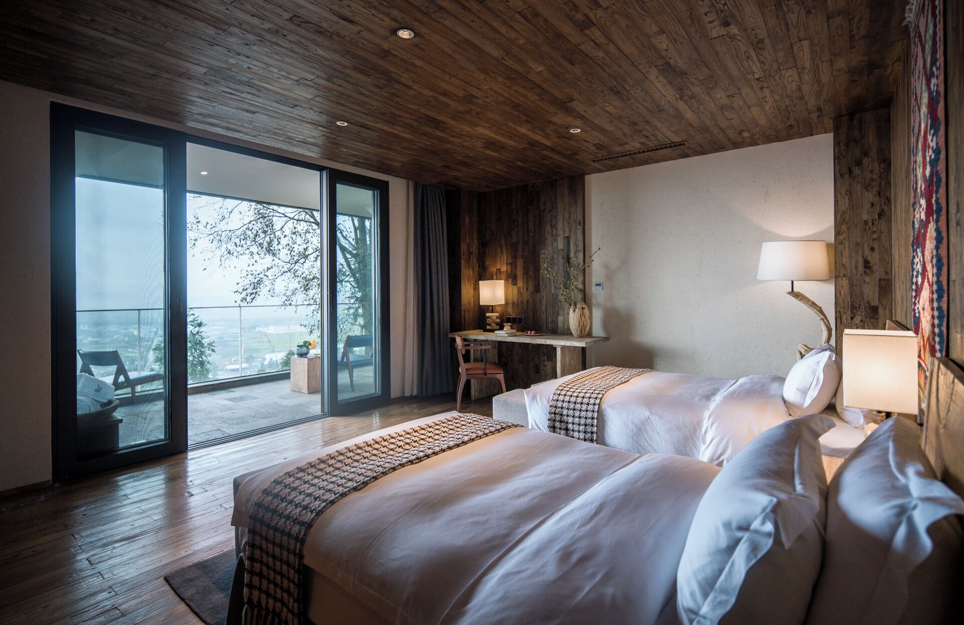 A hotel room with two beds, a desk, a chair, a large window, and a balcony.