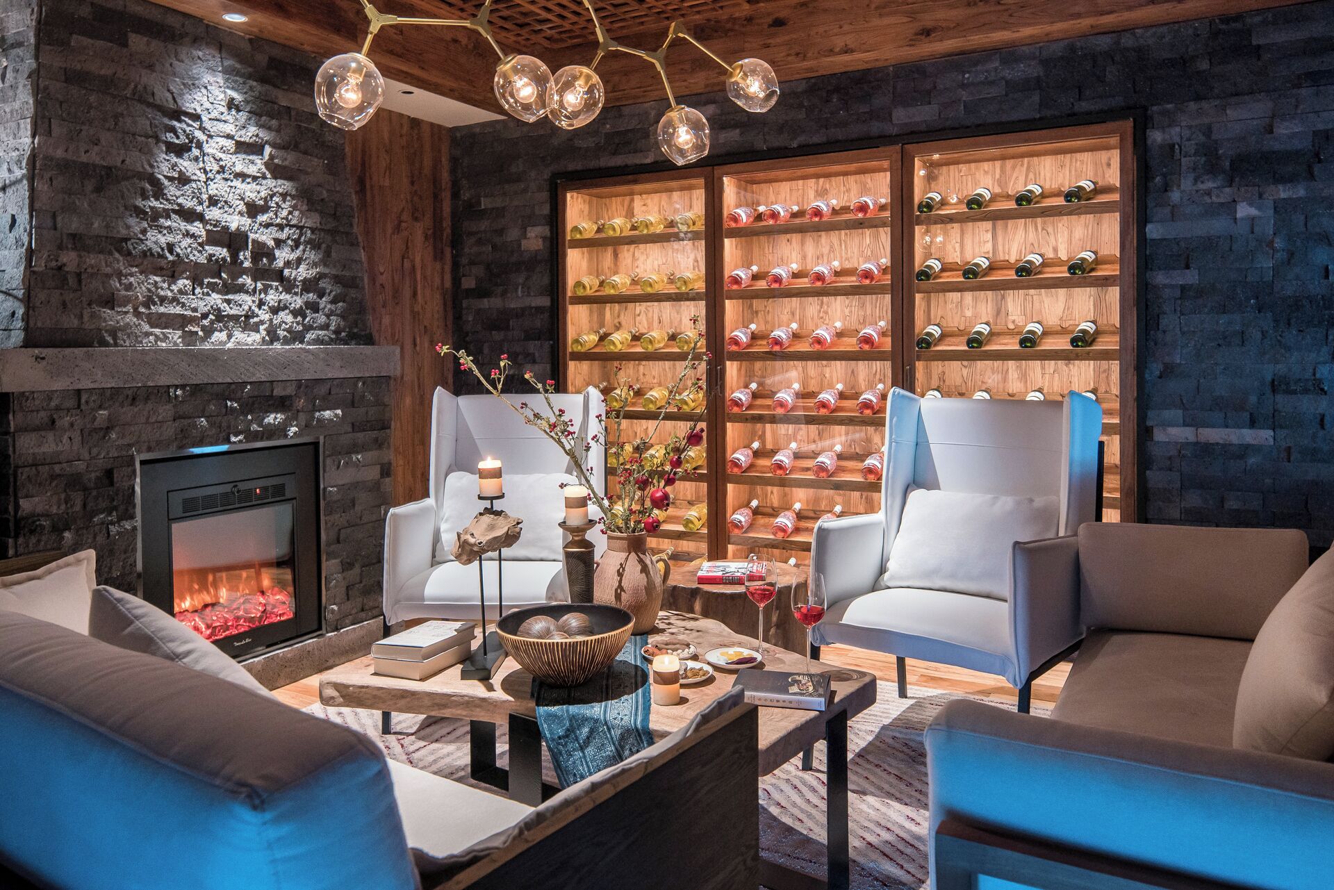 A cozy living room with a fireplace, two armchairs, a coffee table with books and candles, and a wall of wine bottles.