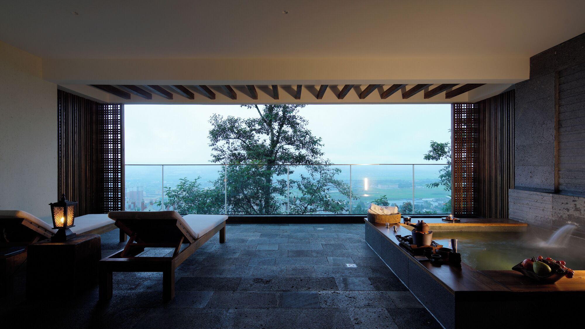 A modern spa room with a large window, a massage table, a wooden bench, and a small table with a teapot and cups.