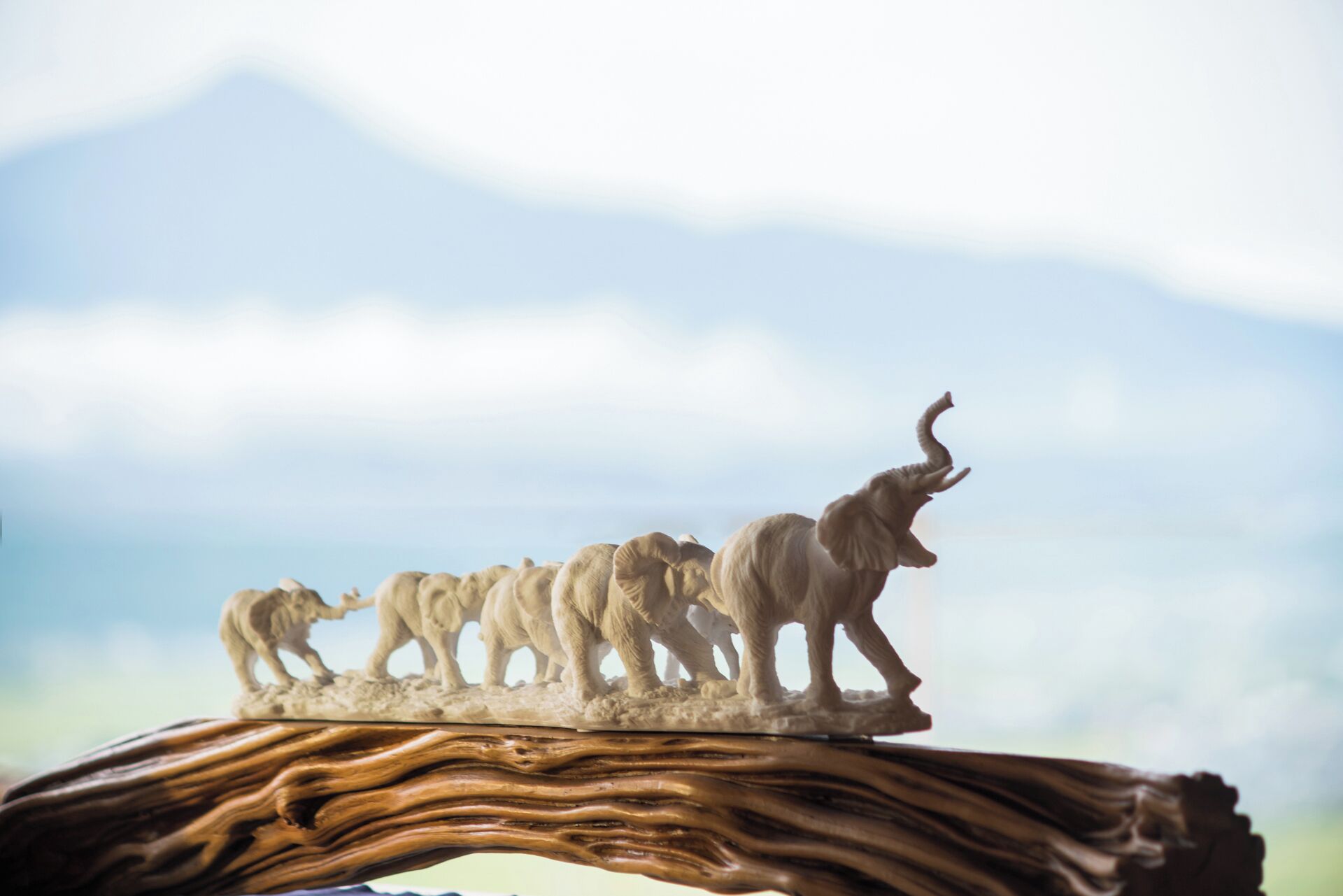 A wooden sculpture of a herd of elephants on a table with a blurred mountain background.