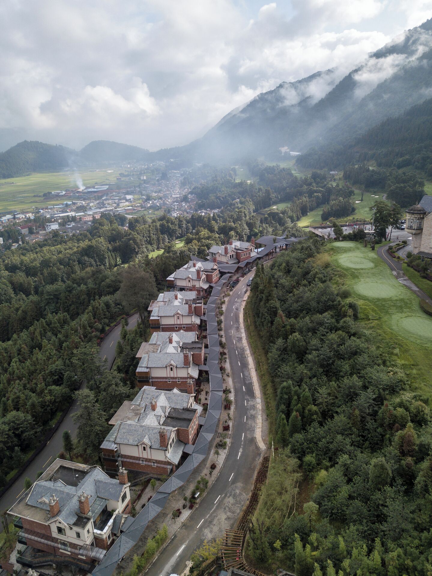 A long row of terraced houses along a winding road, surrounded by dense forest and mountains in the background.
