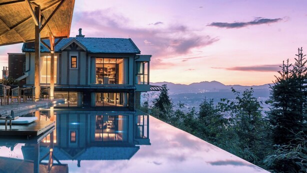Una casa moderna con fachada de vidrio, terraza de madera y una piscina que refleja el entorno.