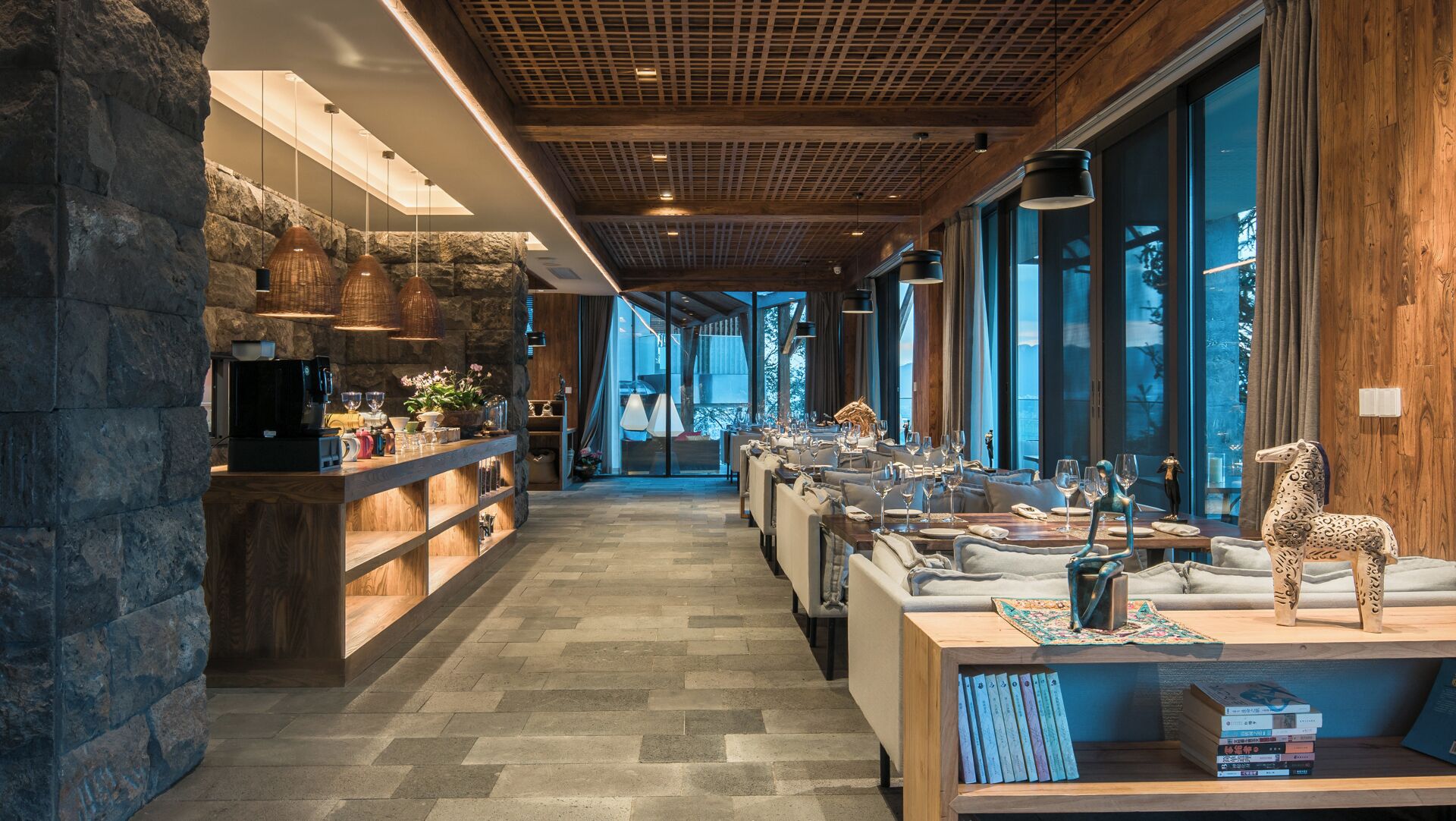 Una hall d'hotel moderna con parete in pietra, soffitto in legno e una lunga zona pranzo con tavoli e sedie.