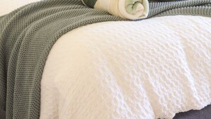 Una cama bien hecha con una manta verde y blanca a rayas, un plaid gris tejido y una almohada blanca con textura.
