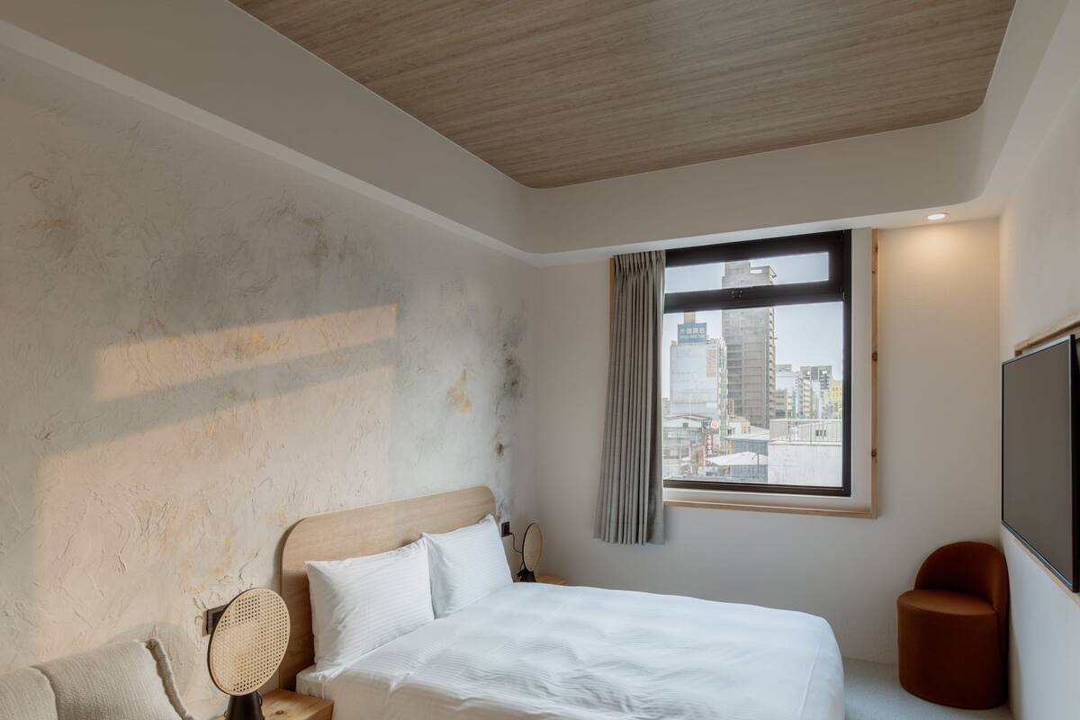 A hotel room with a bed, a wooden ceiling, a window with a city view, a TV, and a small chair.