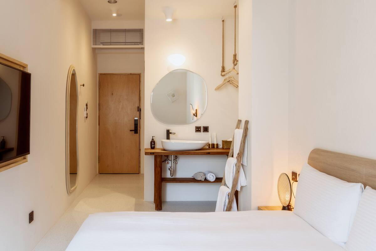 A modern hotel room with a large bed, a wooden headboard, a round mirror, a wooden vanity with a sink, and a door leading to another room.
