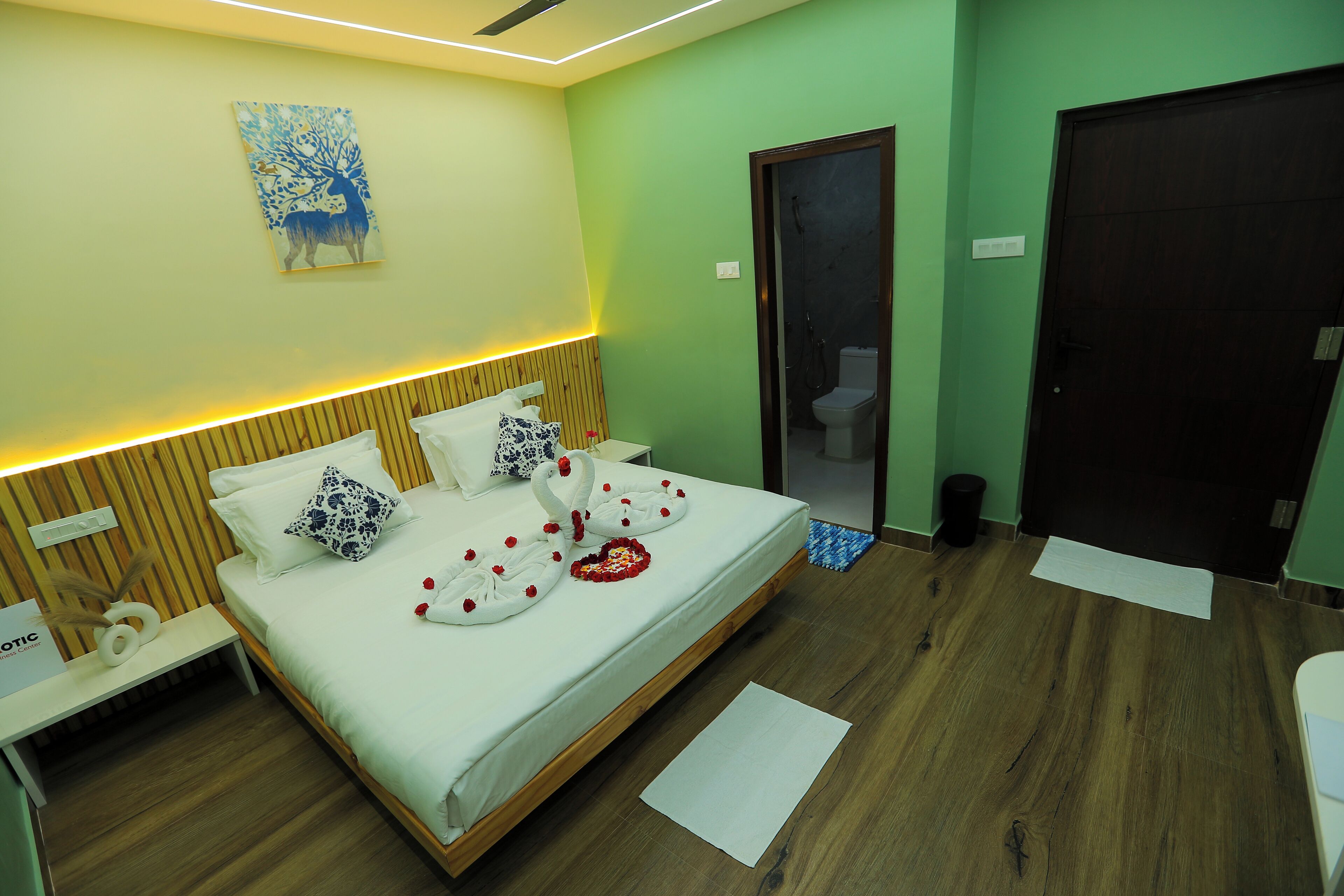 A modern bedroom with a wooden headboard, a bed with white linens and decorative pillows, a bedside table with a lamp, and a painting on the wall.