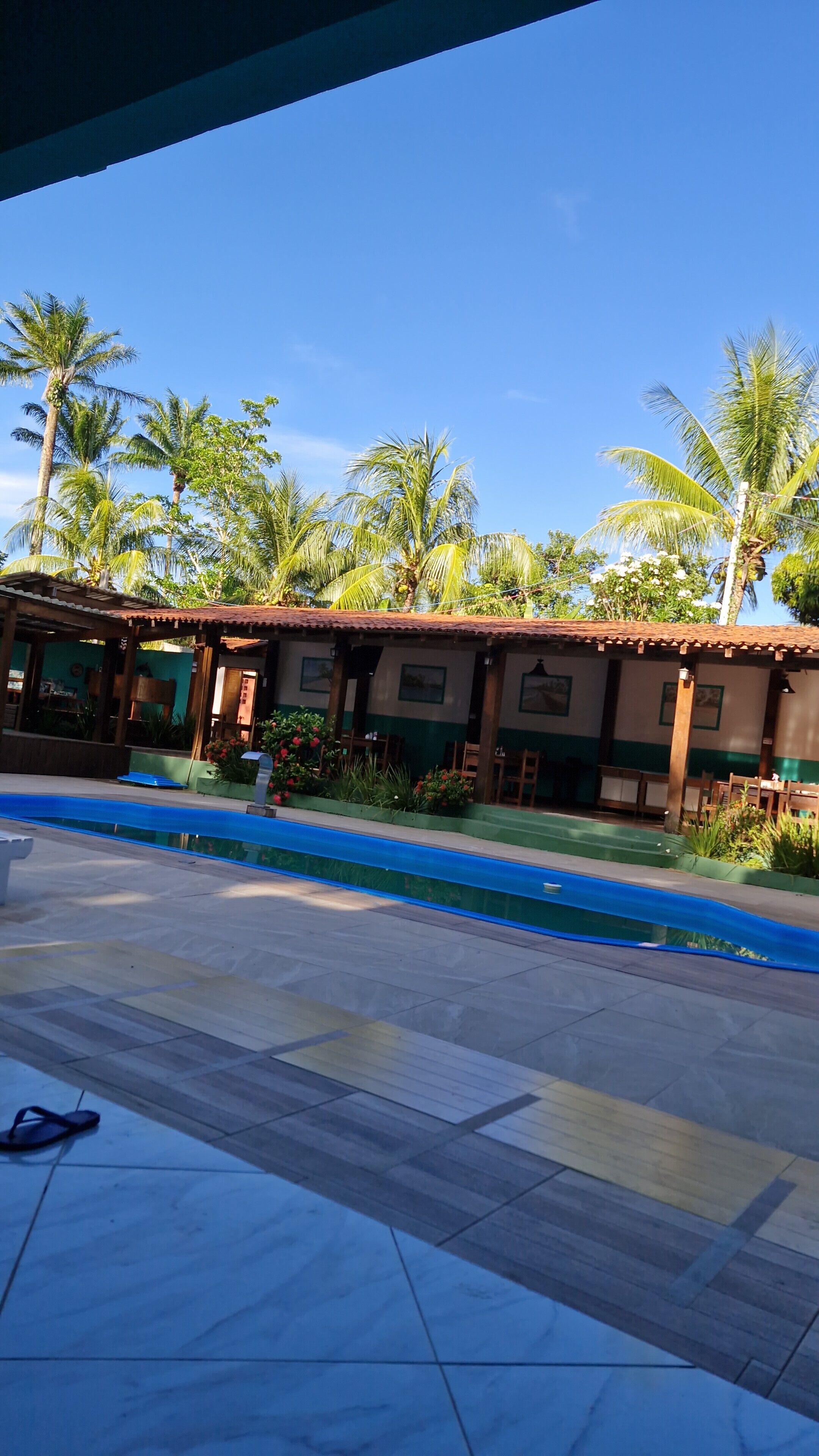 Un área junto a la piscina con suelo de baldosas, una zona de estar cubierta con mesas y sillas, y palmeras al fondo.