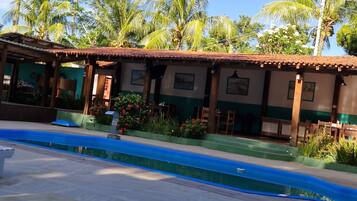 Un área junto a la piscina con suelo de baldosas, una zona de estar cubierta con mesas y sillas, y palmeras al fondo.