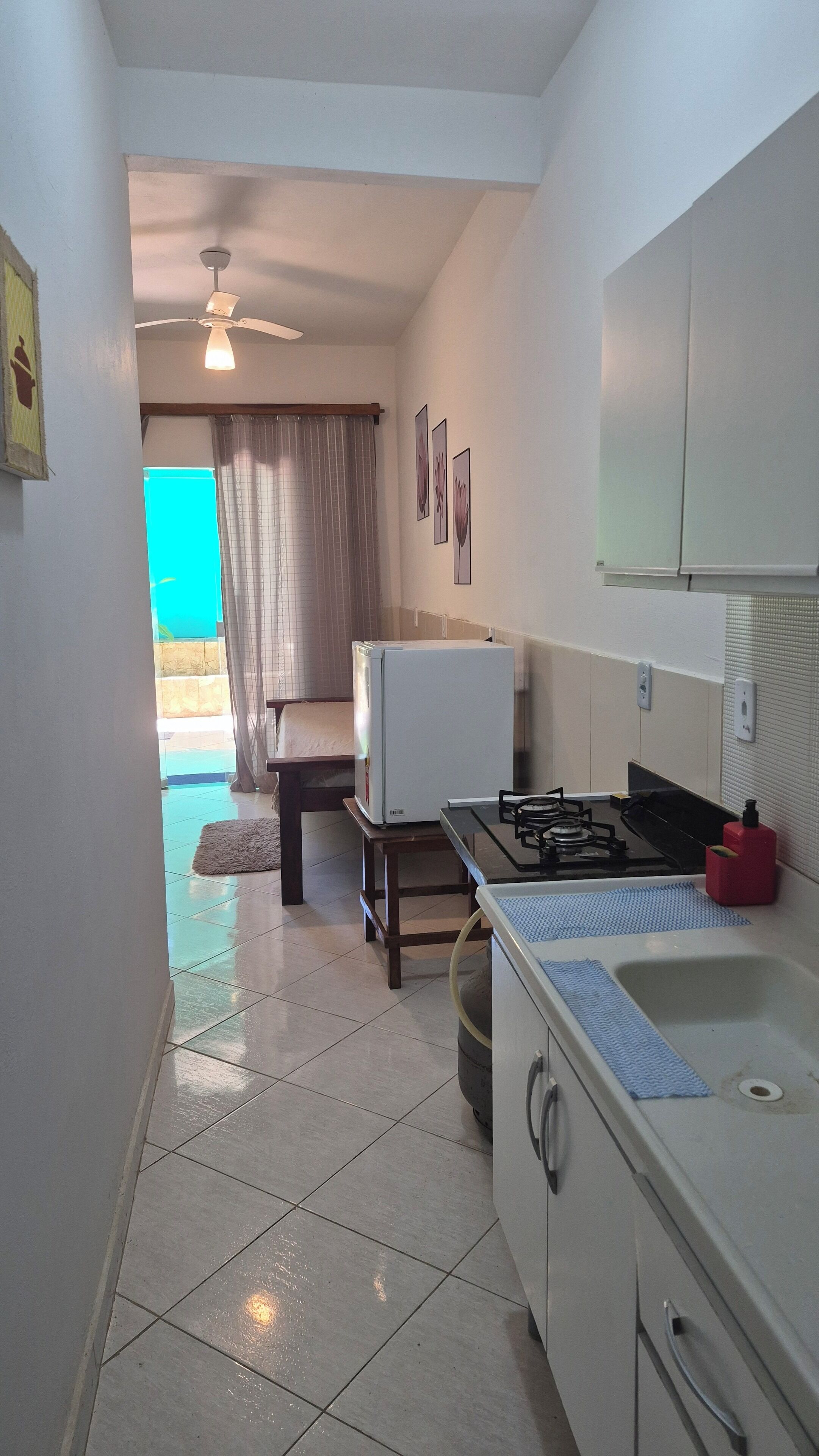 A compact kitchen with a gas stove, a refrigerator, and a sink. There is a ceiling fan and a window with curtains.