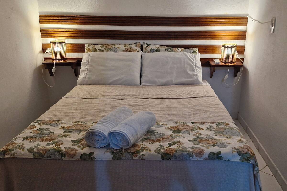 A neatly made bed with a floral bedspread and two rolled towels, a wooden headboard, bedside lamps, and a power outlet on the wall.