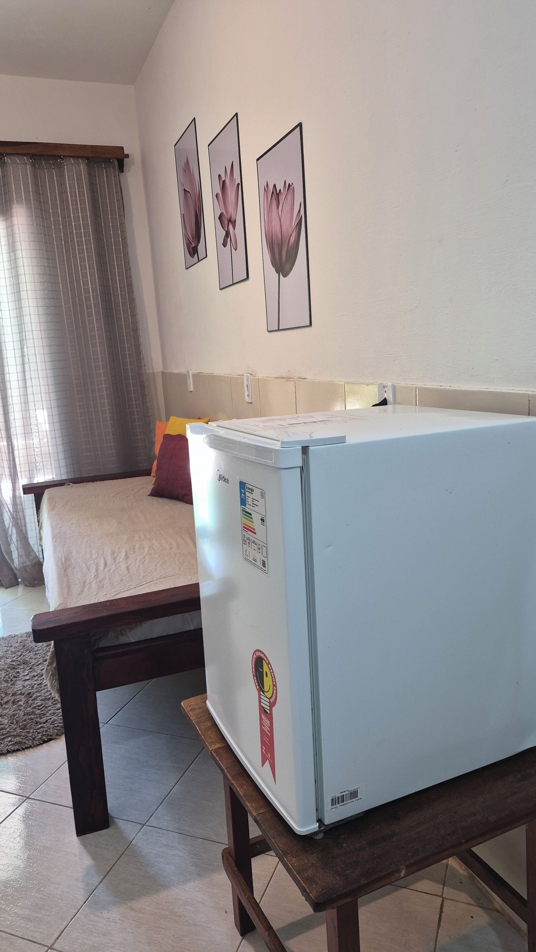 Una habitación con una cama, una mesita y un refrigerador. Hay tres cuadros en la pared.
