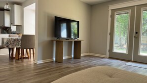 Ein modernes Wohnzimmer mit einem Flachbildfernseher auf einem Holzständer, einem Essbereich mit einer blumigen Tischdecke und einem großen Fenster mit Blick auf Bäume.
