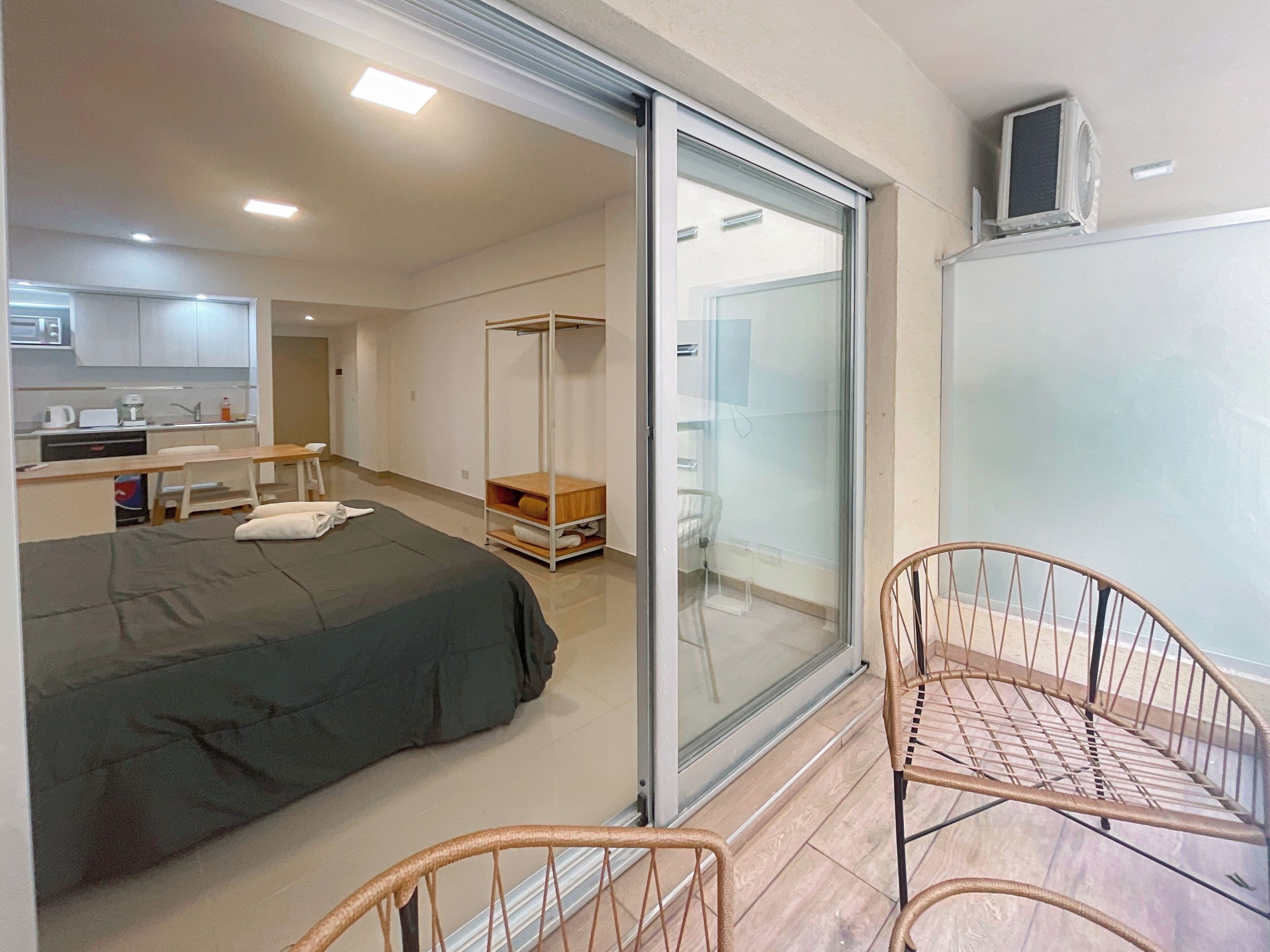 Une chambre d’hôtel compacte comprenant un lit, une petite kitchenette, une porte-fenêtre coulissante donnant sur un balcon et une chaise.