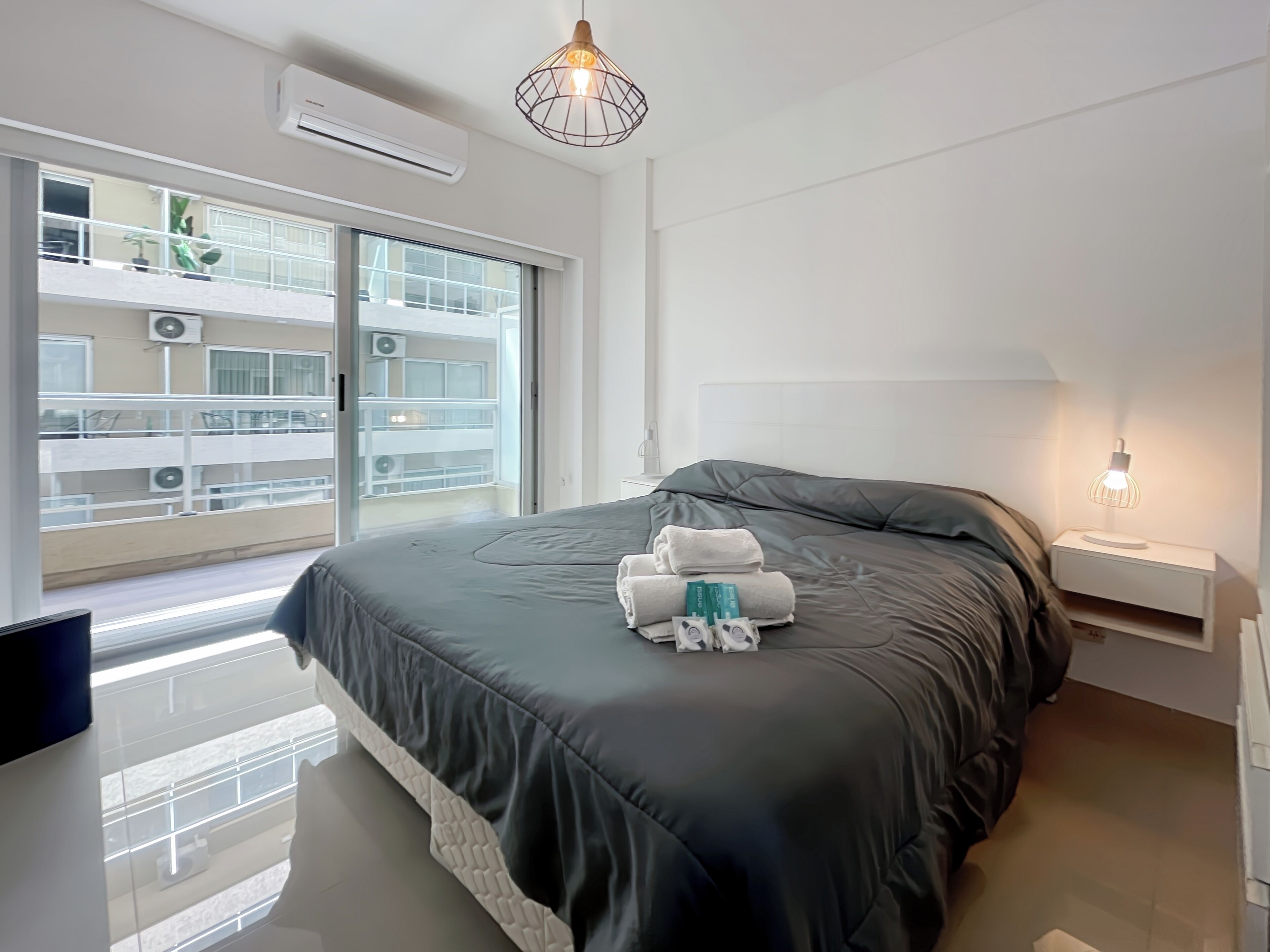 Habitación de hotel moderna con una cama grande, una mesita de noche con lámpara y vistas del exterior del edificio a través de la ventana.