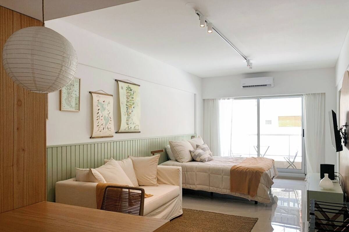 A modern bedroom with a bed, a sofa, and a dining table. There are wall-mounted air conditioning units and a television.