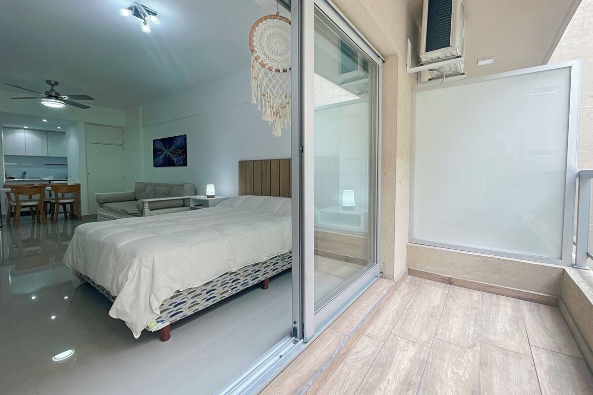 A modern bedroom with a bed, a desk, and a chair. There is a sliding glass door leading to a balcony.
