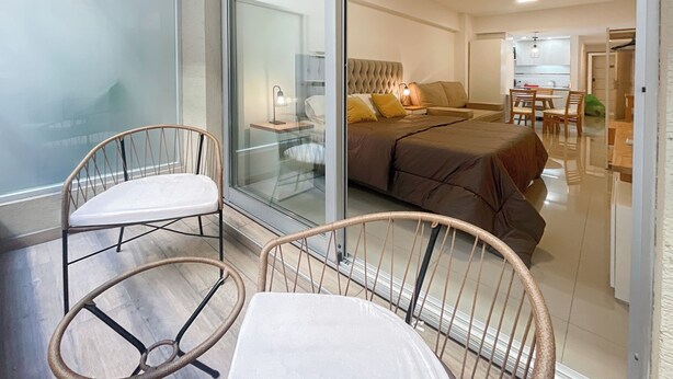 Un dormitorio moderno con una cama grande, una puerta de vidrio que da a un balcón y un área de comedor visible a través del vidrio.