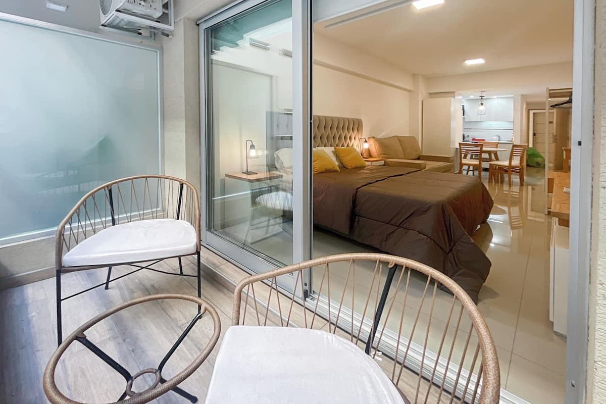 A modern bedroom with a large bed, a glass door leading to a balcony, and a dining area visible through the glass.