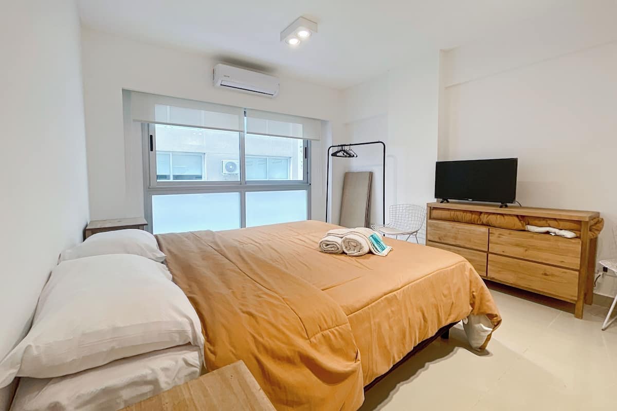 A hotel room with a bed, a TV, a wooden dresser, and a window with a view of buildings.