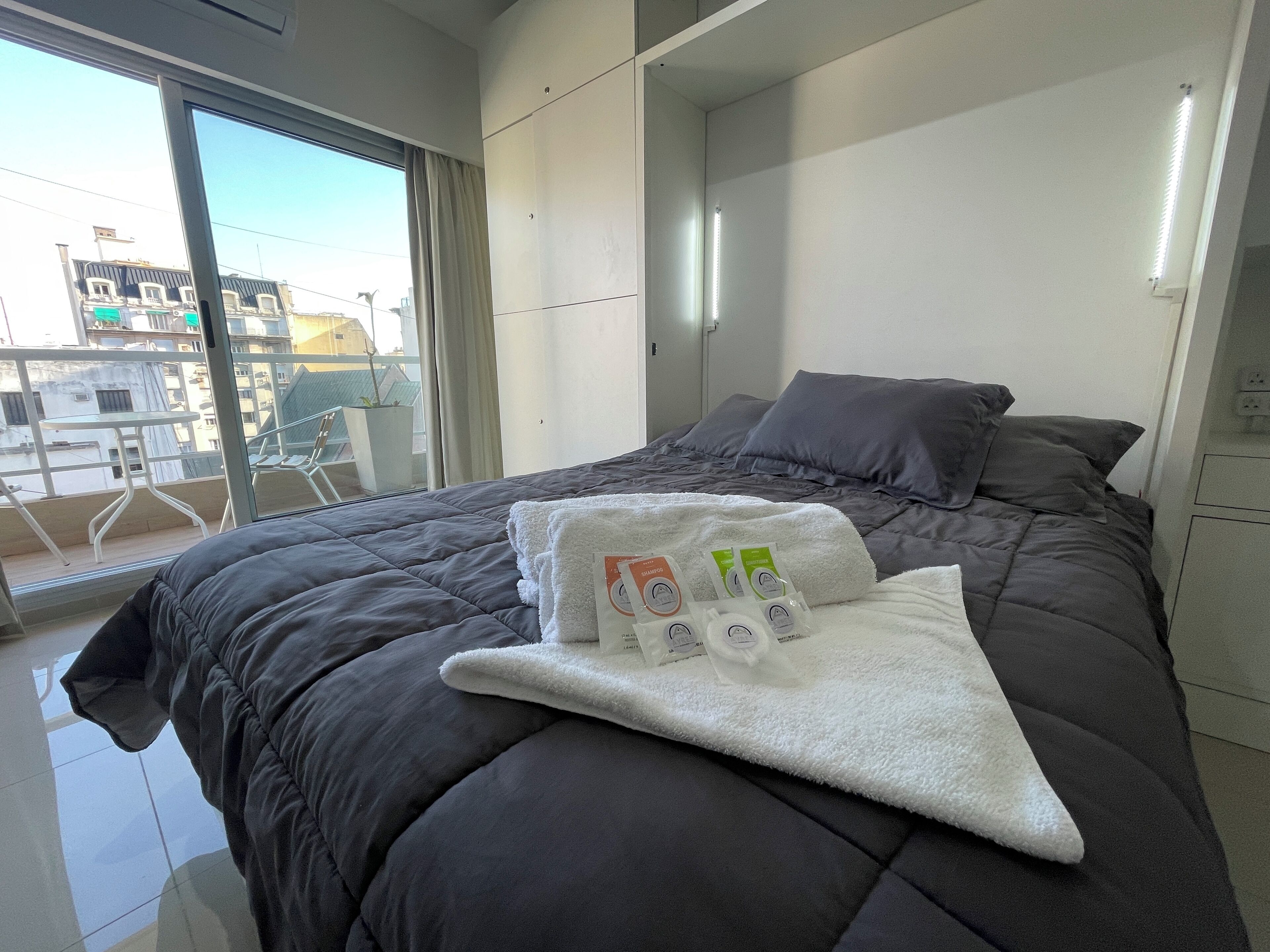 A modern hotel room with a large bed, a view of the city through a large window, and a balcony with a chair.