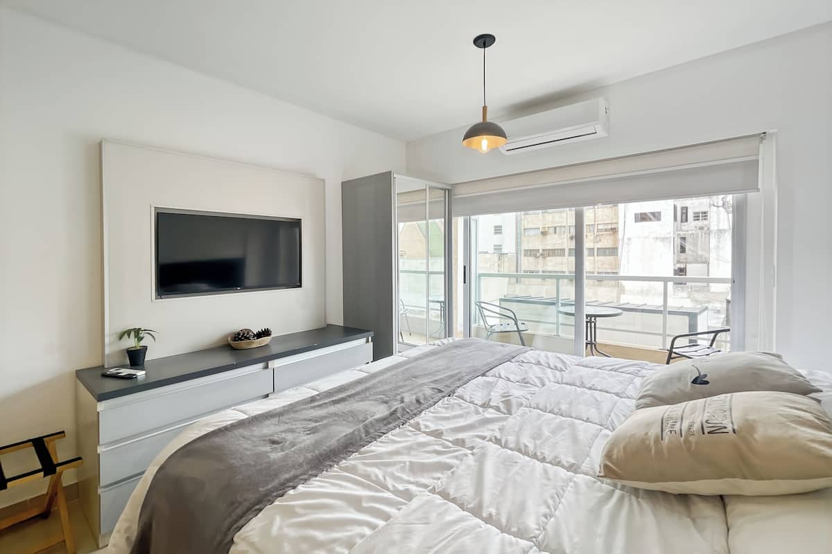 A modern bedroom with a large bed, a TV mounted on the wall, a small desk, and a balcony with a glass railing.