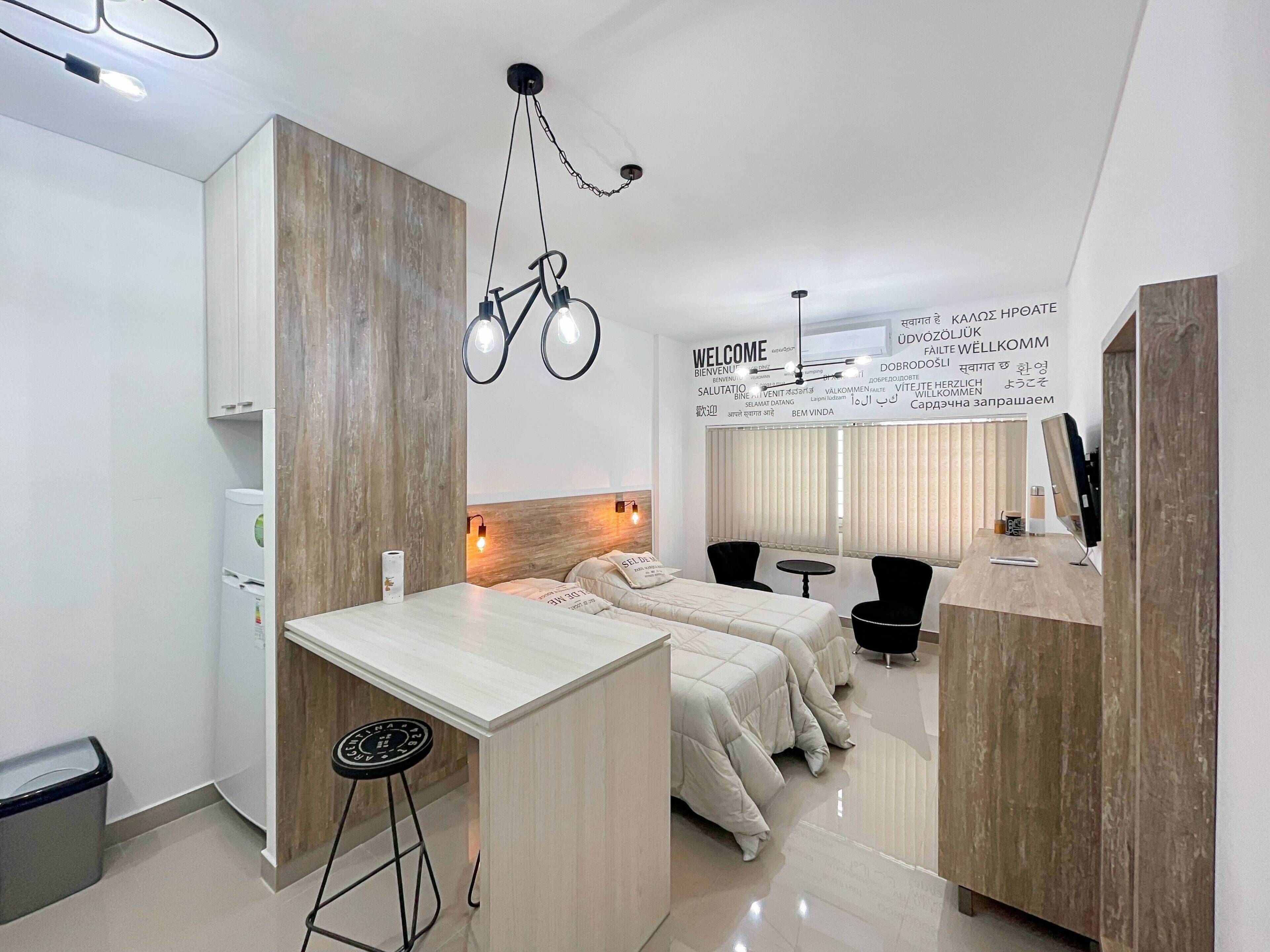 Una habitación de hotel moderna con kitchenette, una cama con cabecera, un escritorio y una silla. Hay un televisor montado en la pared y un cartel con varios idiomas.