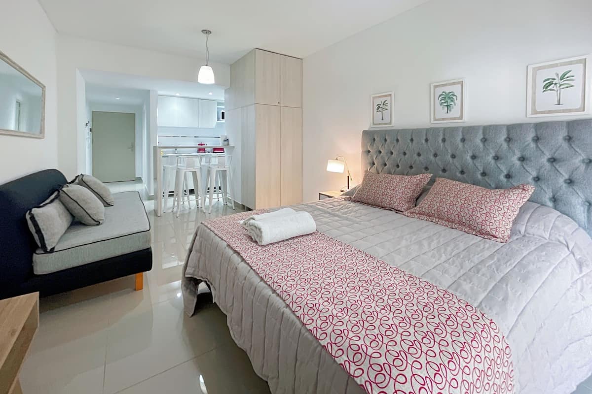 A modern bedroom with a large bed, a sofa, a desk, and a kitchenette in the background.