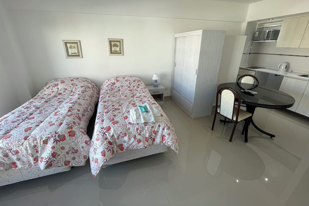 A hotel room with two beds, a small table with a chair, a wardrobe, and a kitchenette.