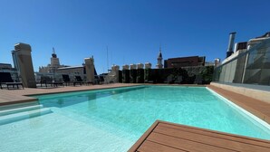 Una piscina en la azotea con agua azul cristalina, rodeada de sillones de descanso y un paisaje urbano de fondo.