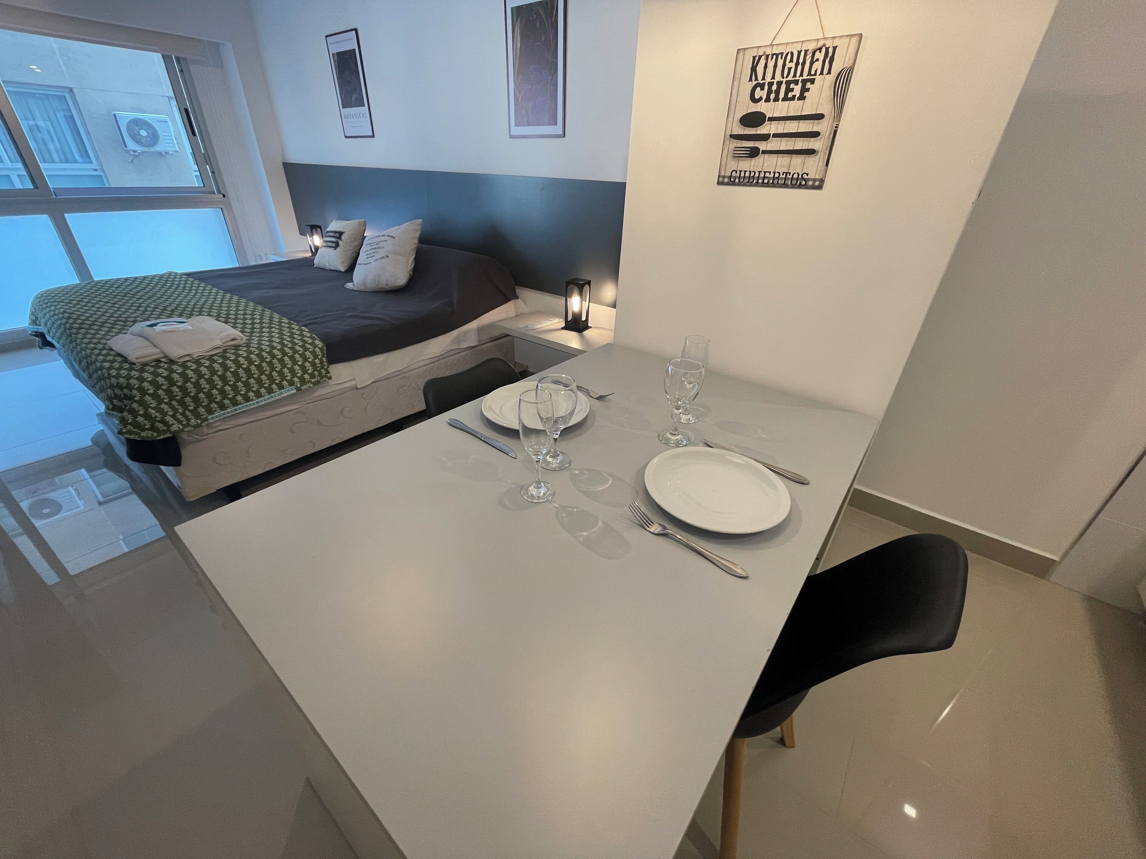 A modern hotel room with a dining area, a bed with a green patterned bedspread, and a kitchen sign on the wall.