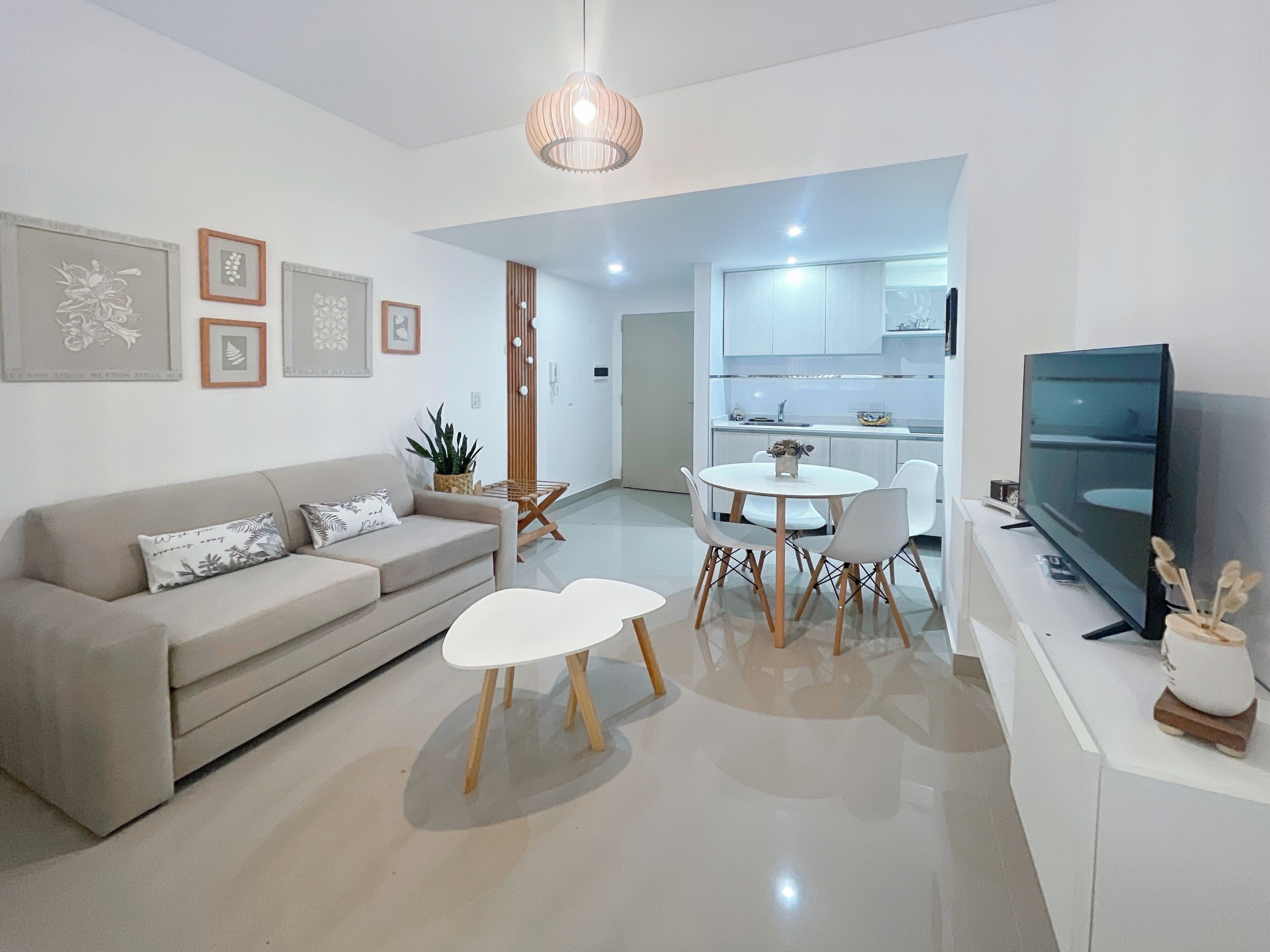 A modern living room with a sofa, coffee table, and dining table. There is a TV on a stand and a kitchen in the background.