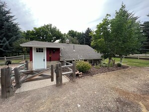 A small building with a red door, surrounded by a wooden fence and a gravel area with plants.