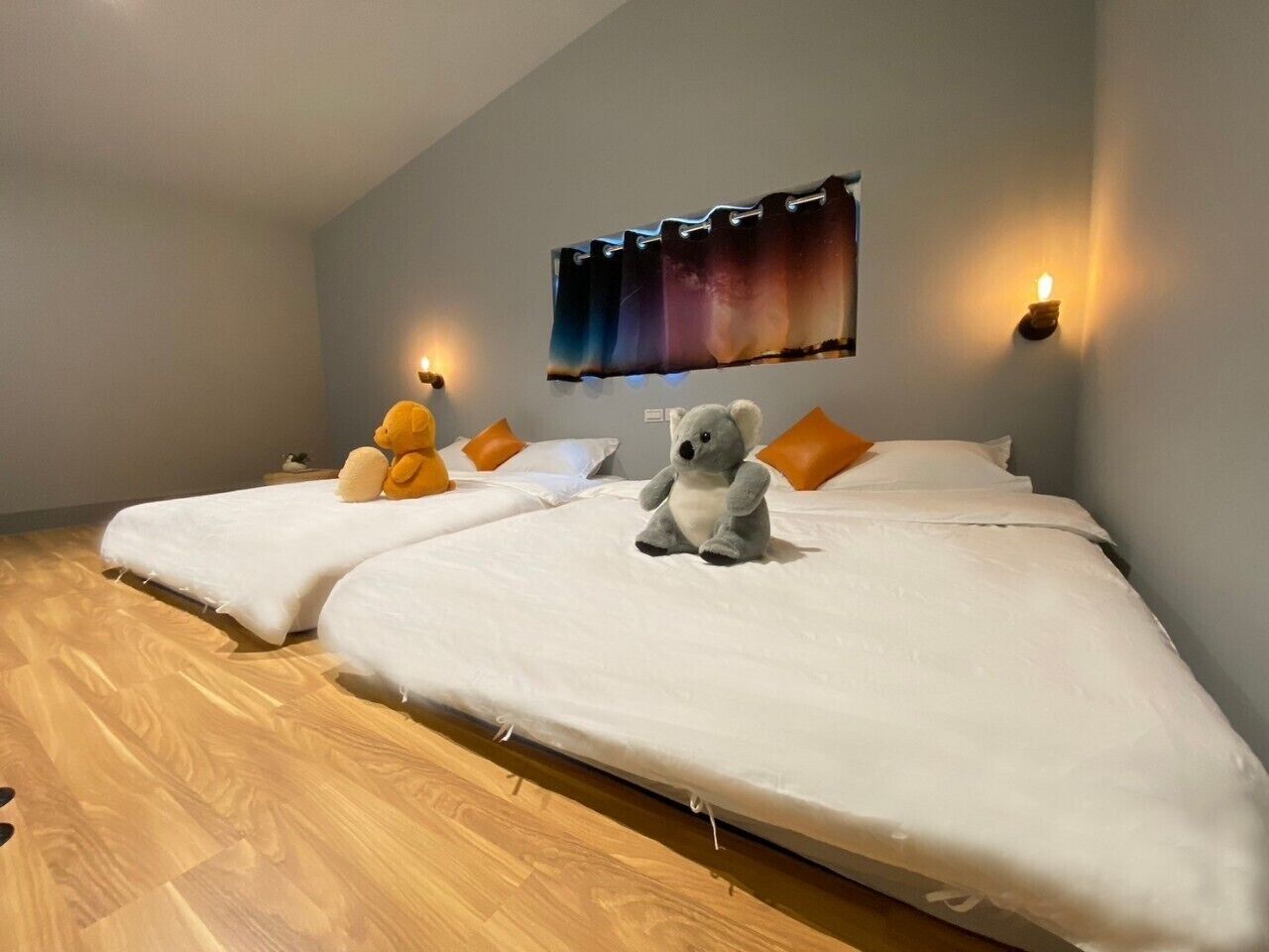 A room with two beds, each with a white mattress and pillows, a wooden floor, and a wall-mounted light fixture.