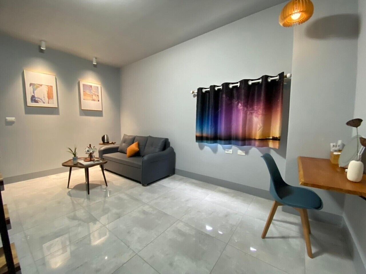 Een moderne woonkamer met een grijze bank, een bijzettafeltje en een blauwe fauteuil. Twee wandlampen en een groot raam met gordijn.