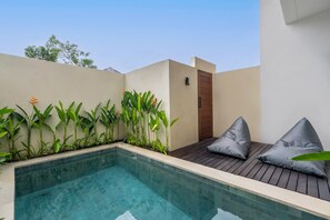 A modern outdoor pool area with a wooden deck and a small seating area with cushions. - Jane Berawa Villa by Betterplace (Canggu)