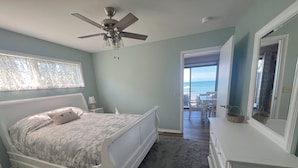A bedroom with a bed, a dresser, a ceiling fan, and a view of the ocean through a door.
