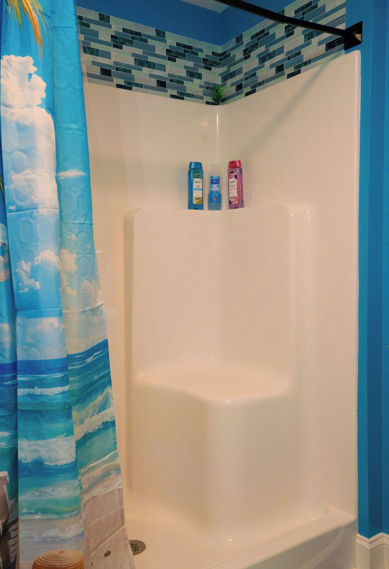 A shower with a blue curtain, a tiled wall, and three bottles of shampoo on the wall.
